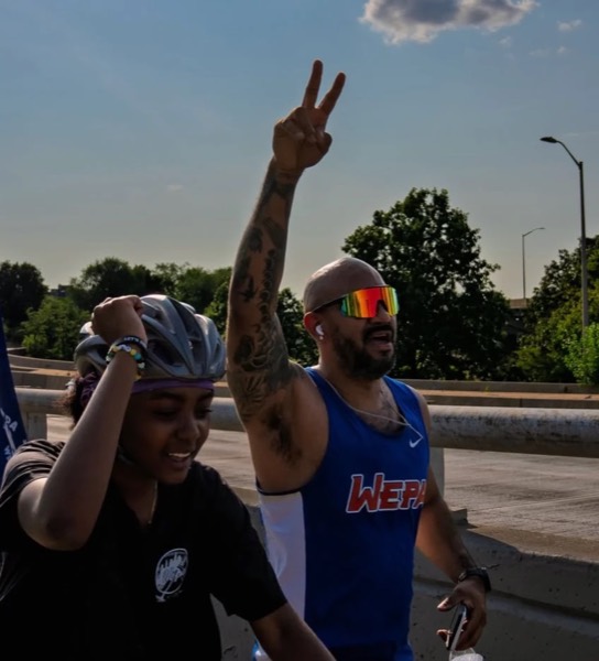 A runner in a WEPA singlet throwing a peace sign beside a cyclist on an overpass at WEPA 9 for 9